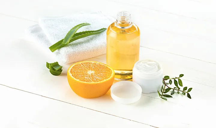 A composition of a towel, aloe leaf, half an orange, a bottle of oil, and a small jar of cream on a light wooden surface.